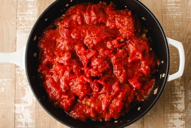 Tomato sauce, diced tomatoes, and water poured over lasagna noodles in a skillet.