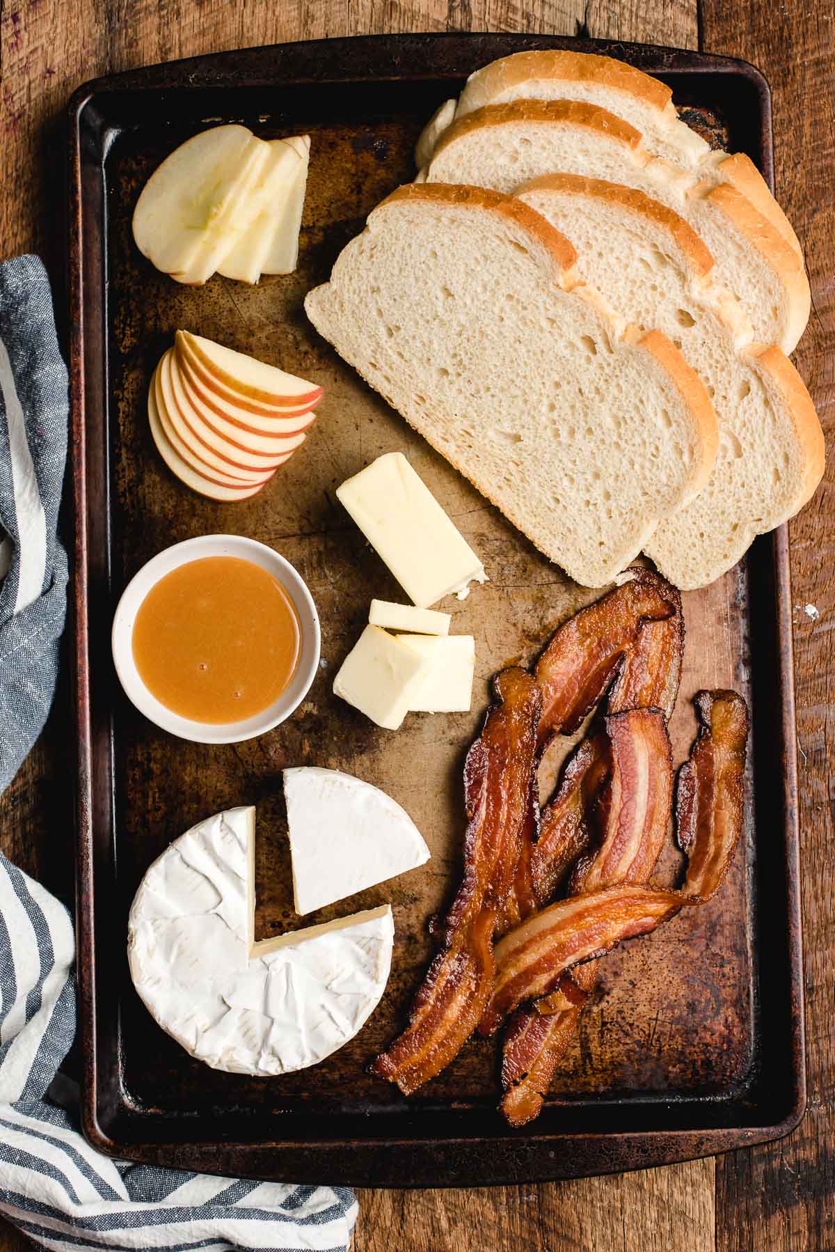 Sheet pan filled with ingredients for grilled cheese: sliced white bread, apple slices, butter, cooked bacon, honey mustard, and a wheel of brie cheese.