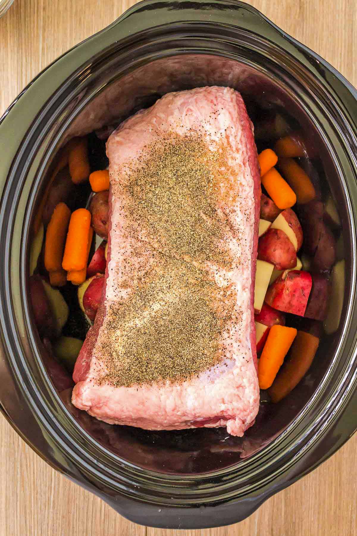 Raw corned beef brisket shown in a crock pot with carrots, potatoes, and seasoning.