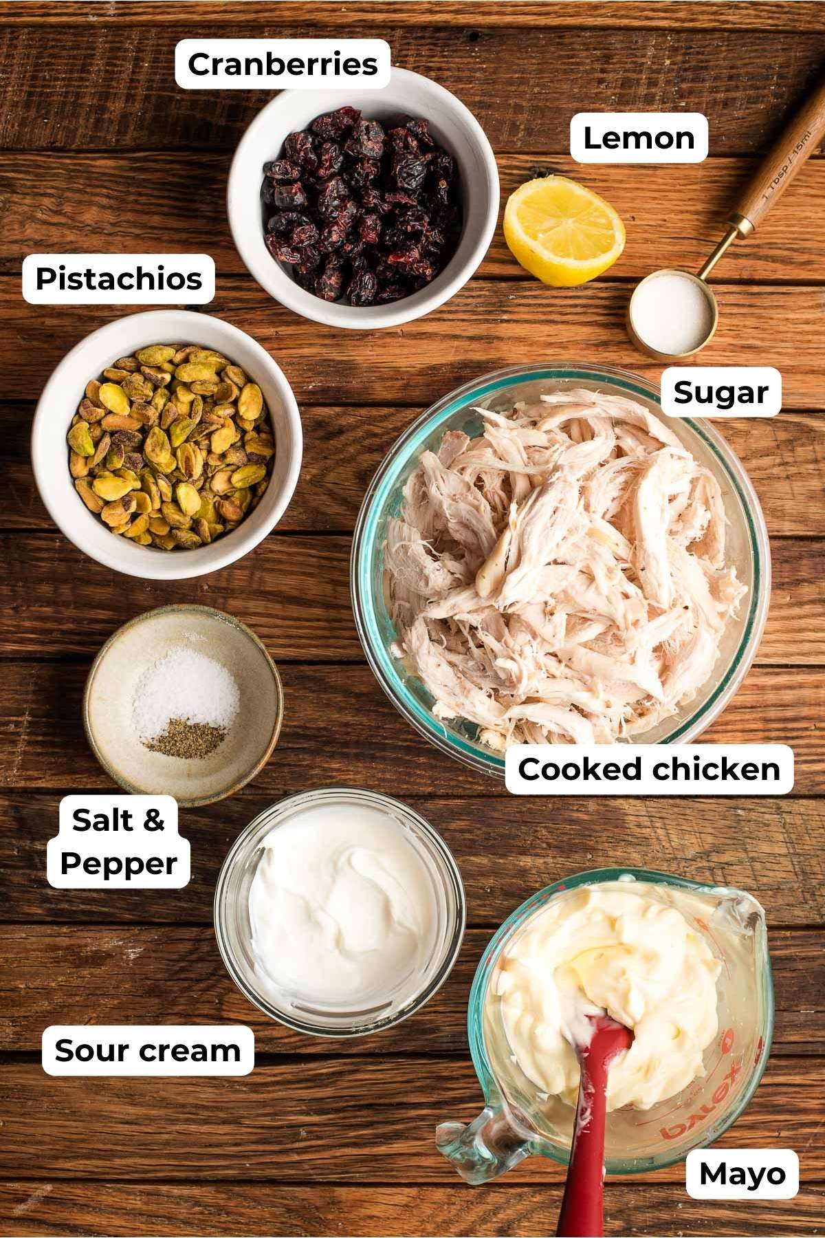 Bowls of dried cranberry, pistachio, salt & pepper, sour cream, mayo, chicken, sugar, and a halved lemon on a wood background.