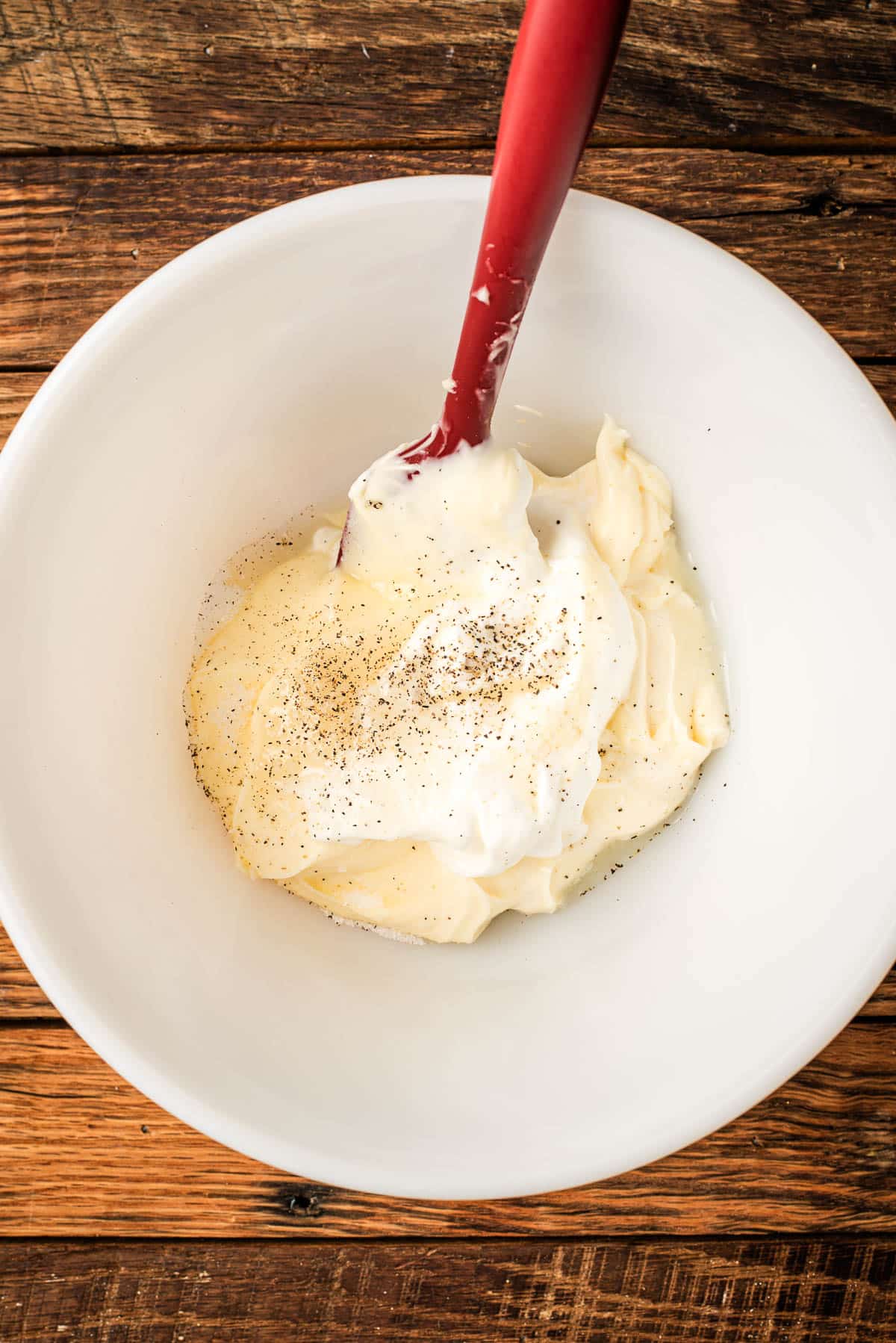 Bowl of mayonnaise, sour cream, sugar, lemon juice, and seasonings with a spatula.
