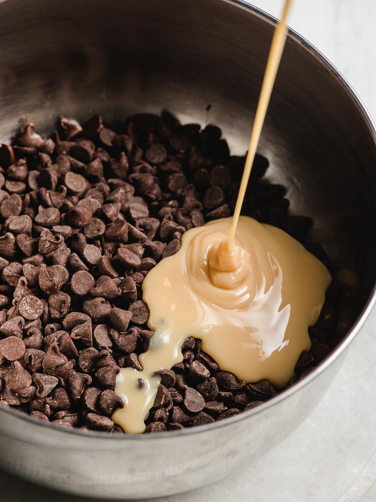 Sweetened condensed milk being poured into a bowl of chocolate chips.