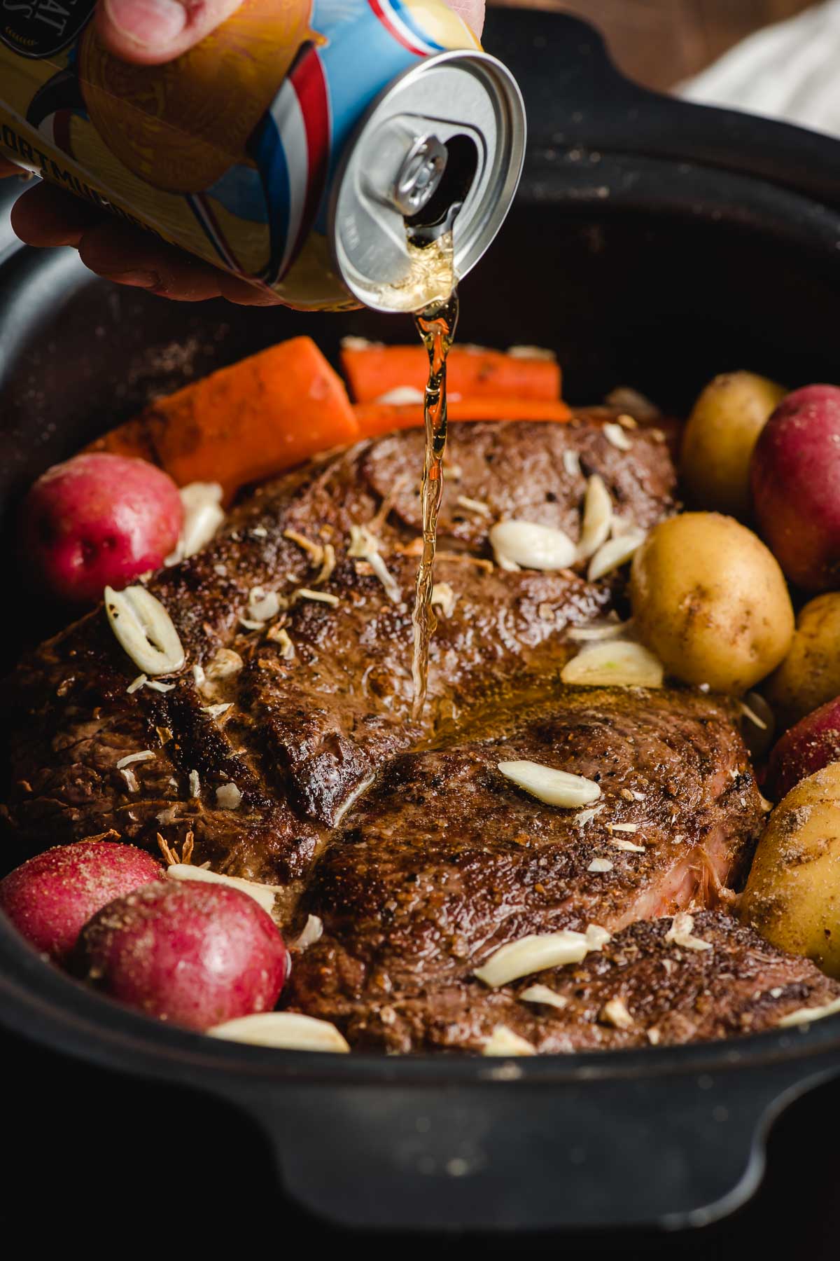 Can of beer being poured over a pot roast in a crock pot.