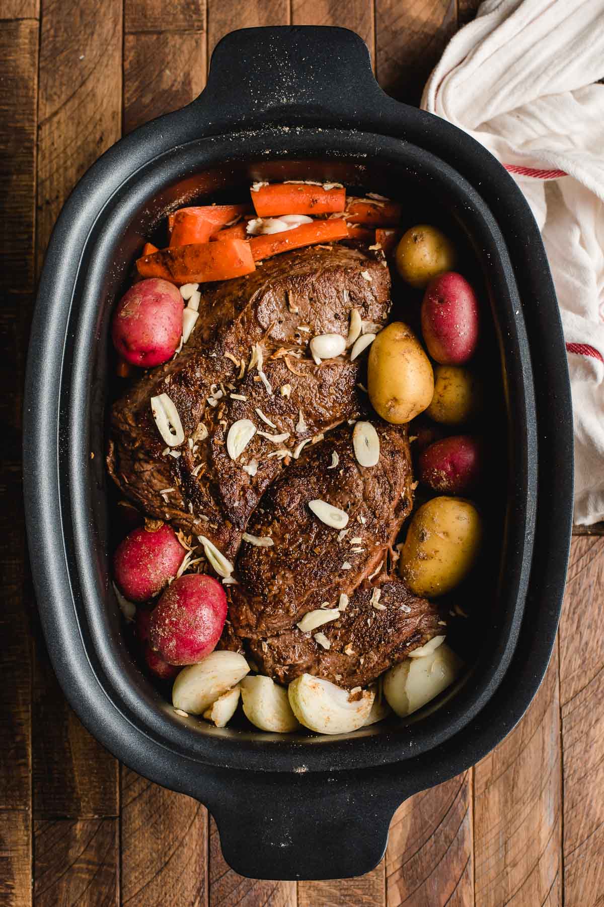 Crock Pot filled with browned pot roast, potatoes, onions, carrots, and garlic.