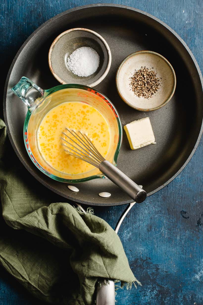 Ingredients for scrambled eggs shown in a skillet- cup of whisked raw egg, pat of butter, salt, and pepper.
