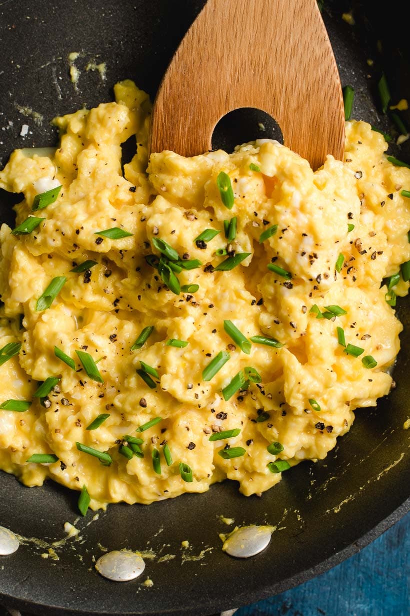 Creamy scrambled eggs showin in a skillet with a wooden spoon scooping them.