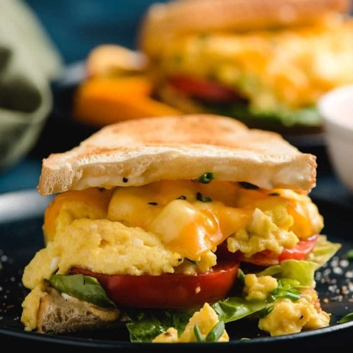 A scrambled egg sandwich seen from the side with layer of lettuce, tomato slices, scrambled eggs, cheese, and toasted bread.