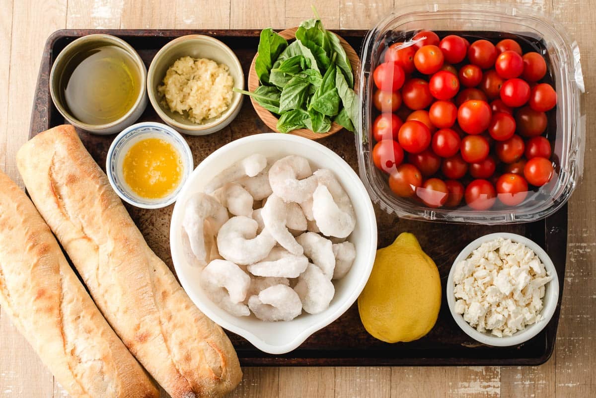 Raw shrimp, garlic, olive oil, lemon juice, feta cheese, cherry tomatoes, and French baguetta on a baking sheet.