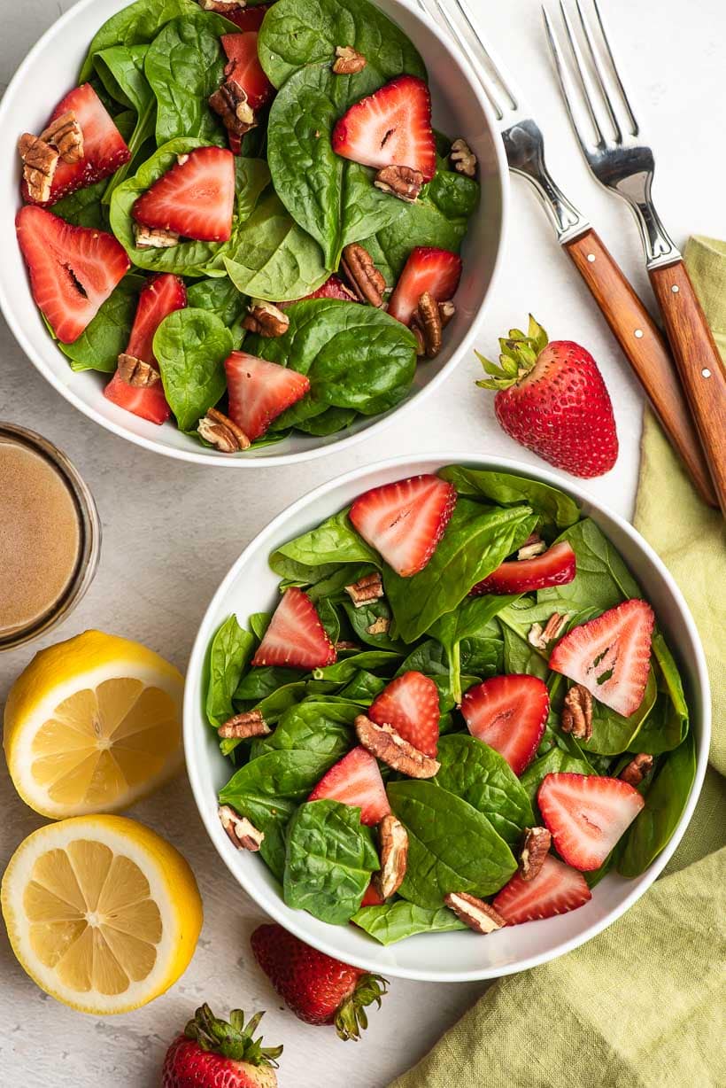 two white bowls filled with spinach salad with strawberries, plus a halved lemon and two forks