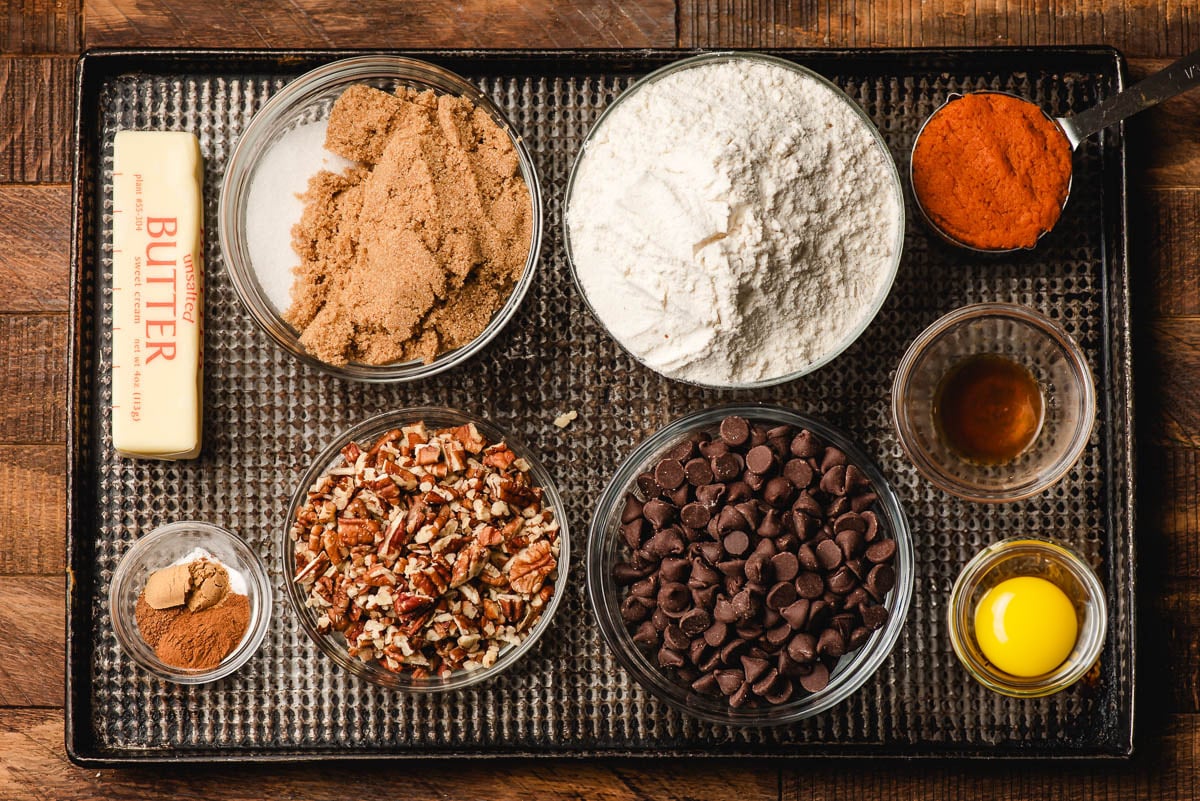 Bowls of flour, spices, pumpkin puree, sugars, butter, pecans, chocolate chips, vanilla, and egg yolks on a baking sheet.