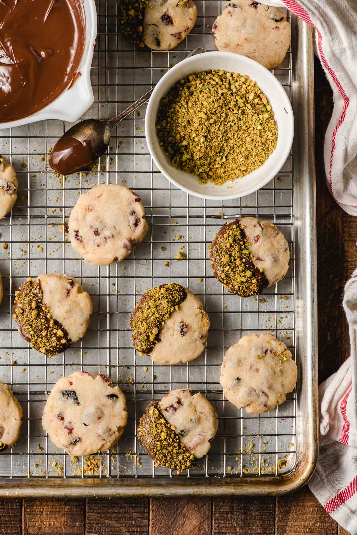 Baking sheet with a cooling rack on top. Multiple cranberry shortbread cookies are on top, some are dipped in semi-sweet chocolate and ground pistachios, others are plain.