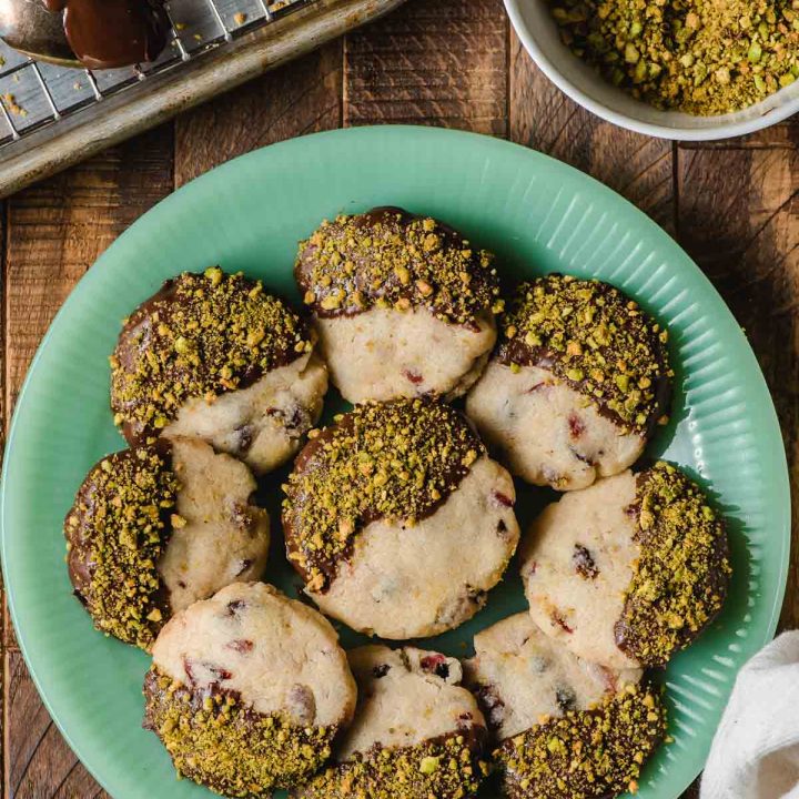 Chocolate dipped cranberry shortbread cookie sprinkled with pistachios and displayed on a green jadette plate.