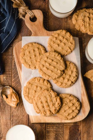 Chewy peanut butter cookies on a wood cutting board surrounded by glasses of milk and a spoonful of peanut butter.