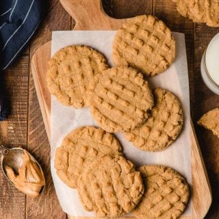 Chewy peanut butter cookies on a wood cutting board surrounded by glasses of milk and a spoonful of peanut butter.