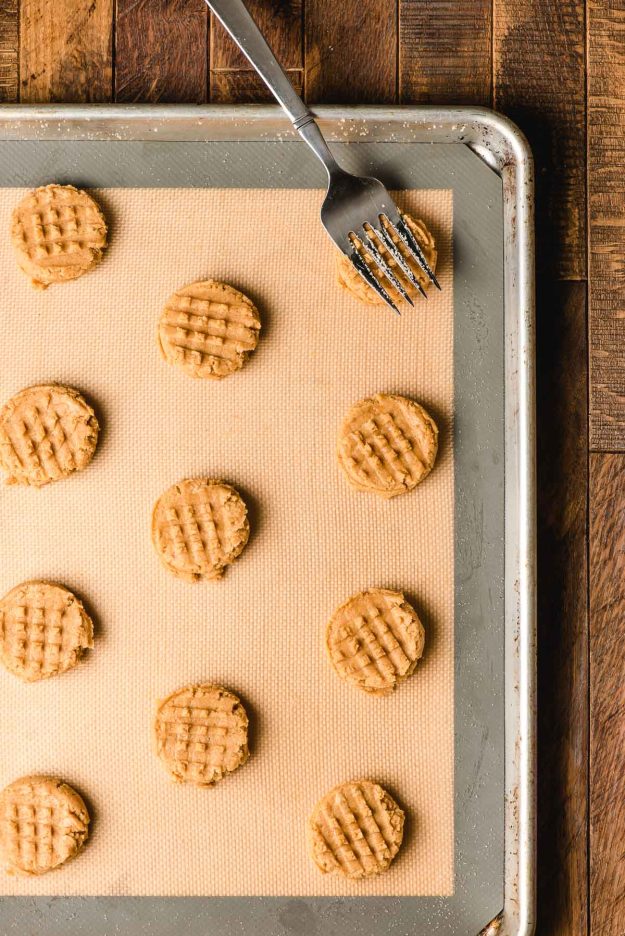 A big serving fork pressing a waffle pattern on the top of a tray of unbaked peanut butter cookies.