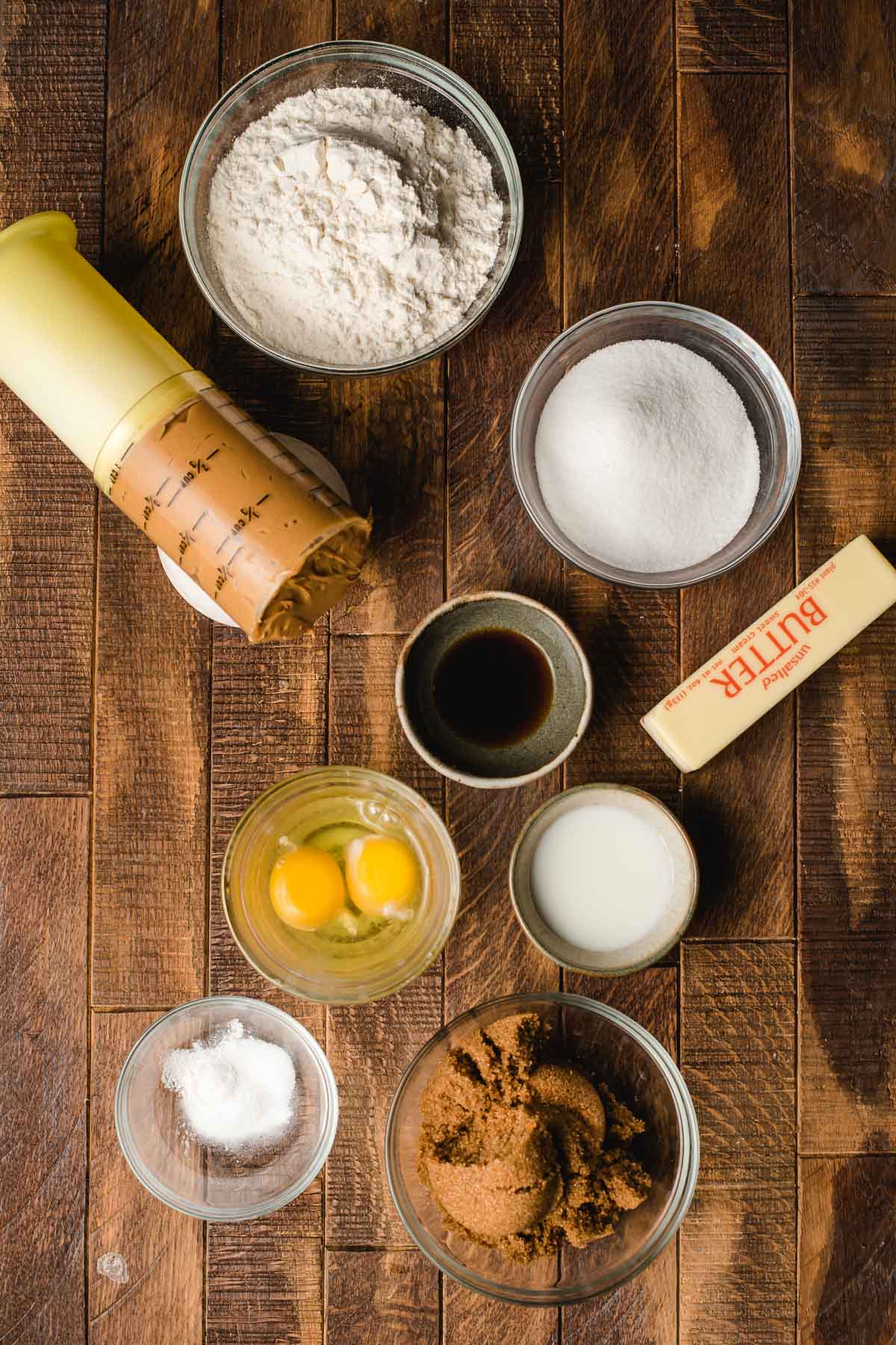 Baking ingredients in small bowls, including peanut butter, butter, eggs, sugar, flour, vanilla, salt, and baking soda.