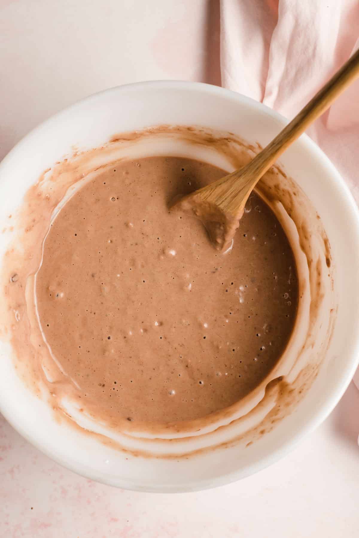 Chocolate waffle batter being stirred by a wooden spoon in a white bowl.