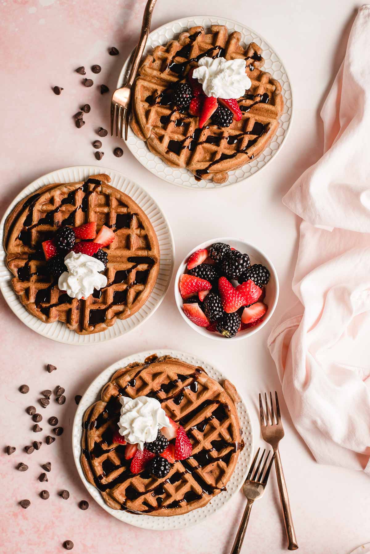 Three white plates each holding a waffle with chocolate, berries, and whipped cream, on a pink background with chocolate chips scattered throughout.