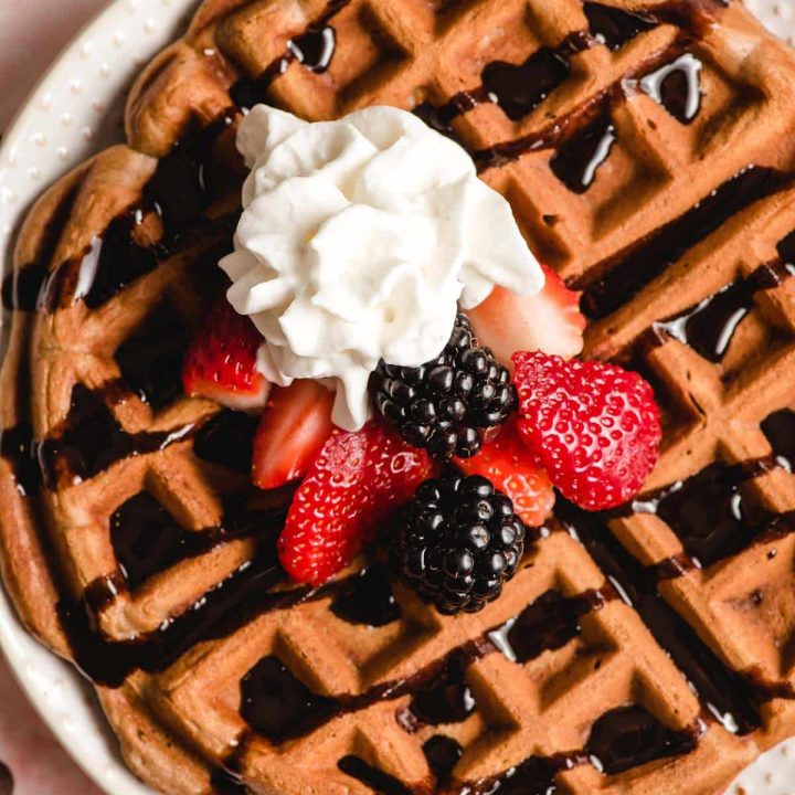 Overhead image of a double chocolate waffle with chocolate syrup, sliced strawberries, blackberries, and whipped cream.