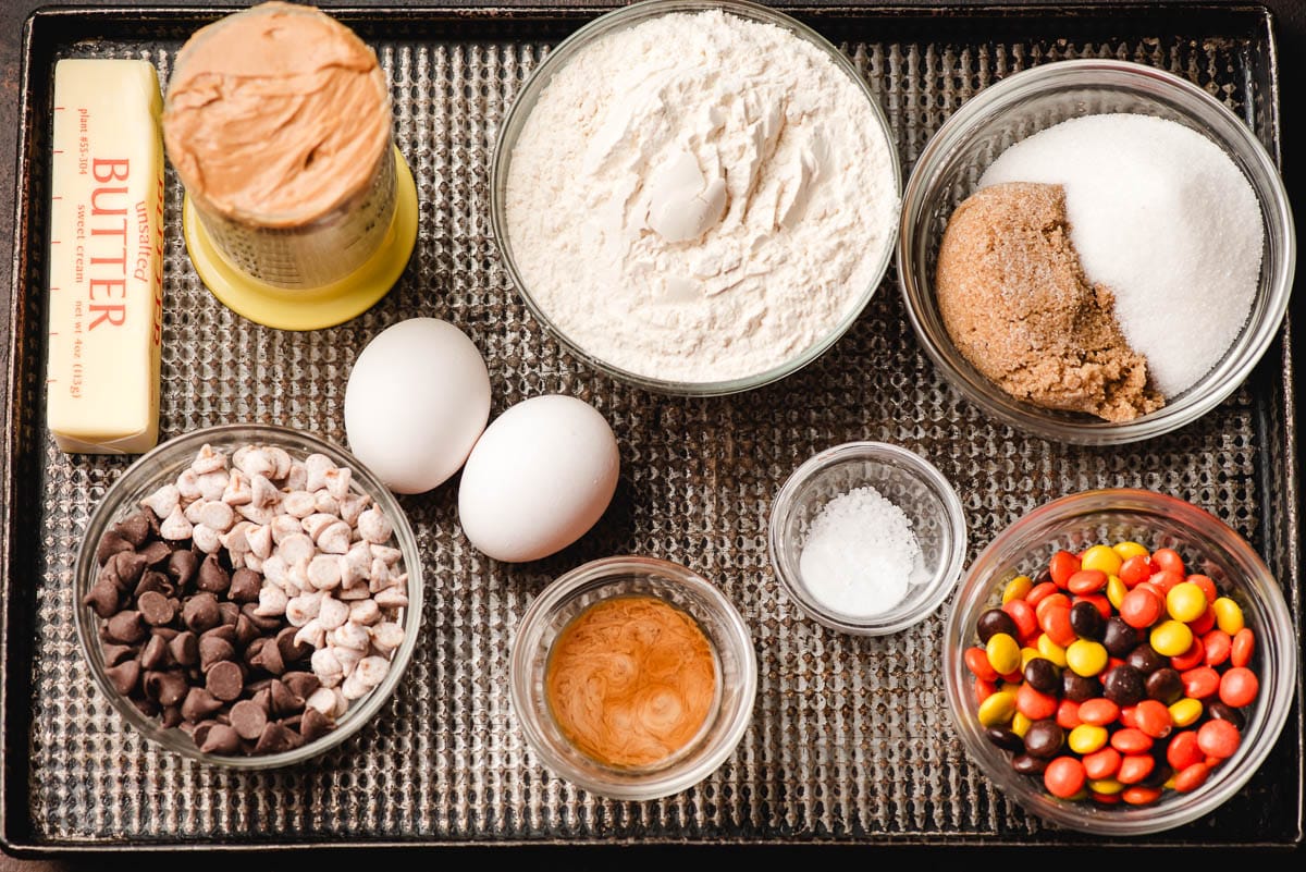 Peanut butter, stick of butter, chocolate chips, peanut butter chips, eggs, flour, sugars, salt, baking soda, vanilla, and Reese's pieces on a sheet pan.