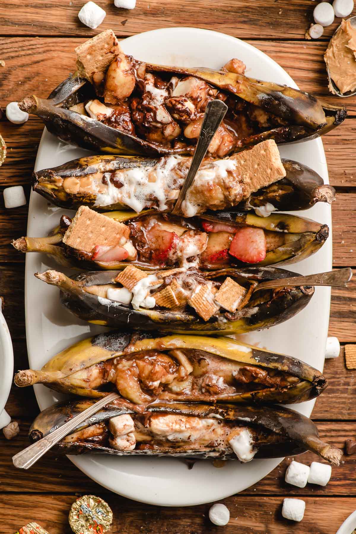 Six campfire banana boats, each stuffed with different toppings, inclduing chocoalte chips, strawberries, marshmallows, and graham crackers.