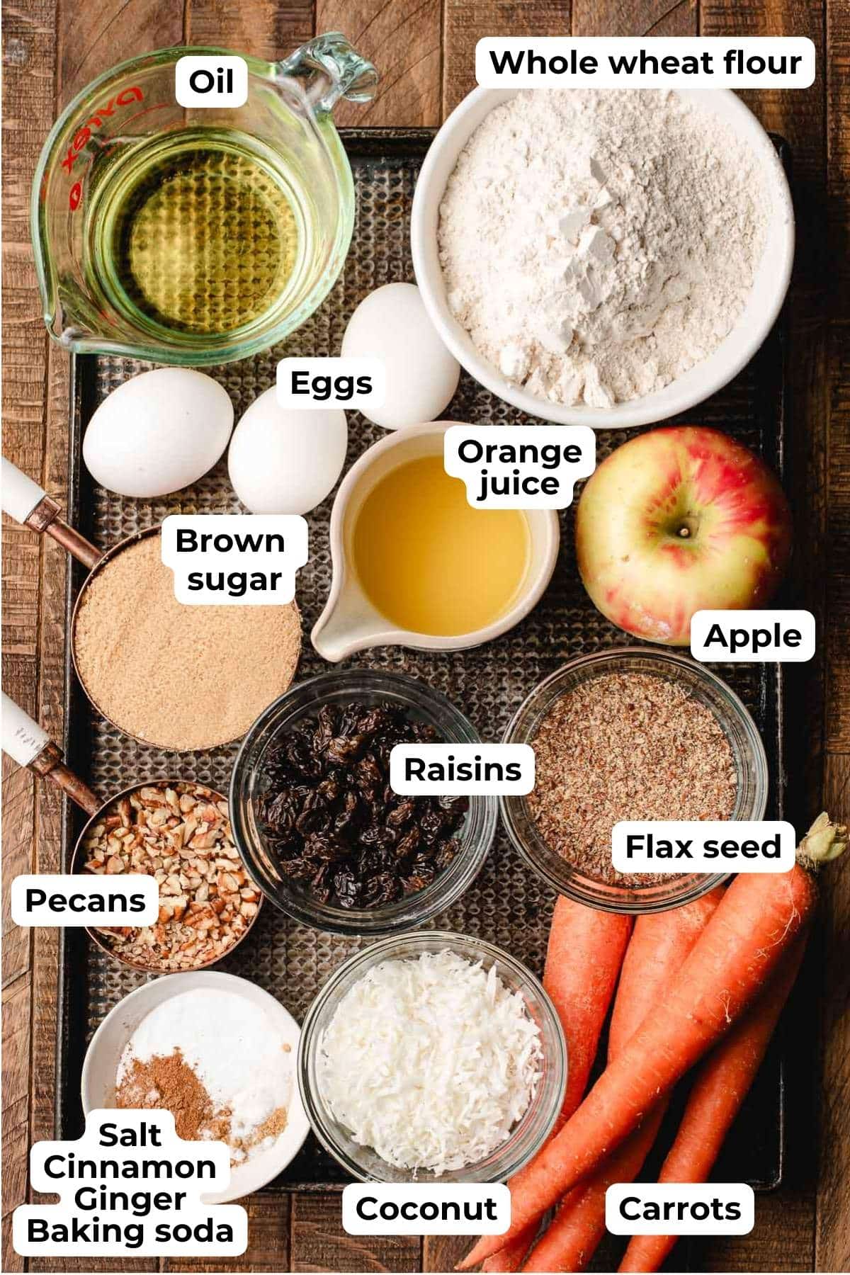 Flour, apple carrot, coconut, raisins, pecans, spices, brown sugar, orange juice, eggs, and oil on a wood background.