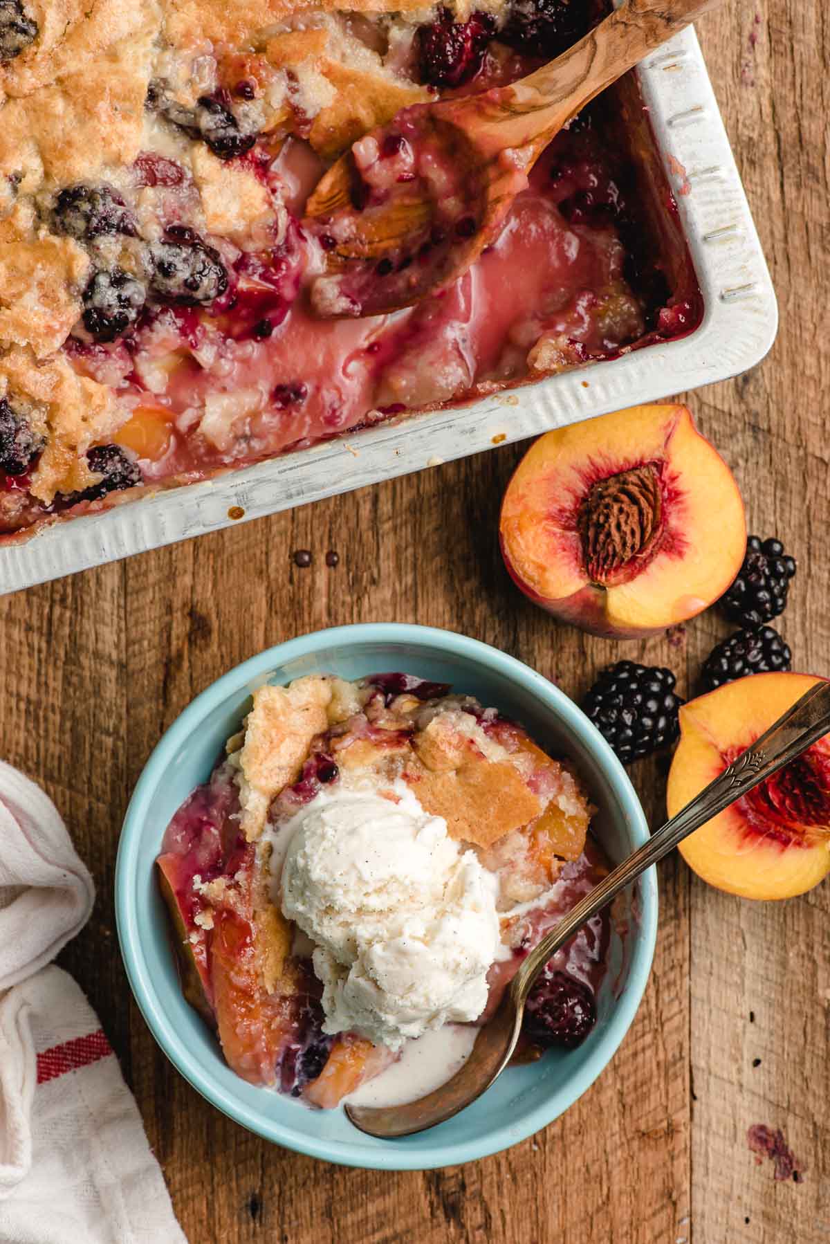 Pan of blackberry peach cobbler with a wooden serving spoon, slantingly a trencher with a scoop of cobbler and ice cream.