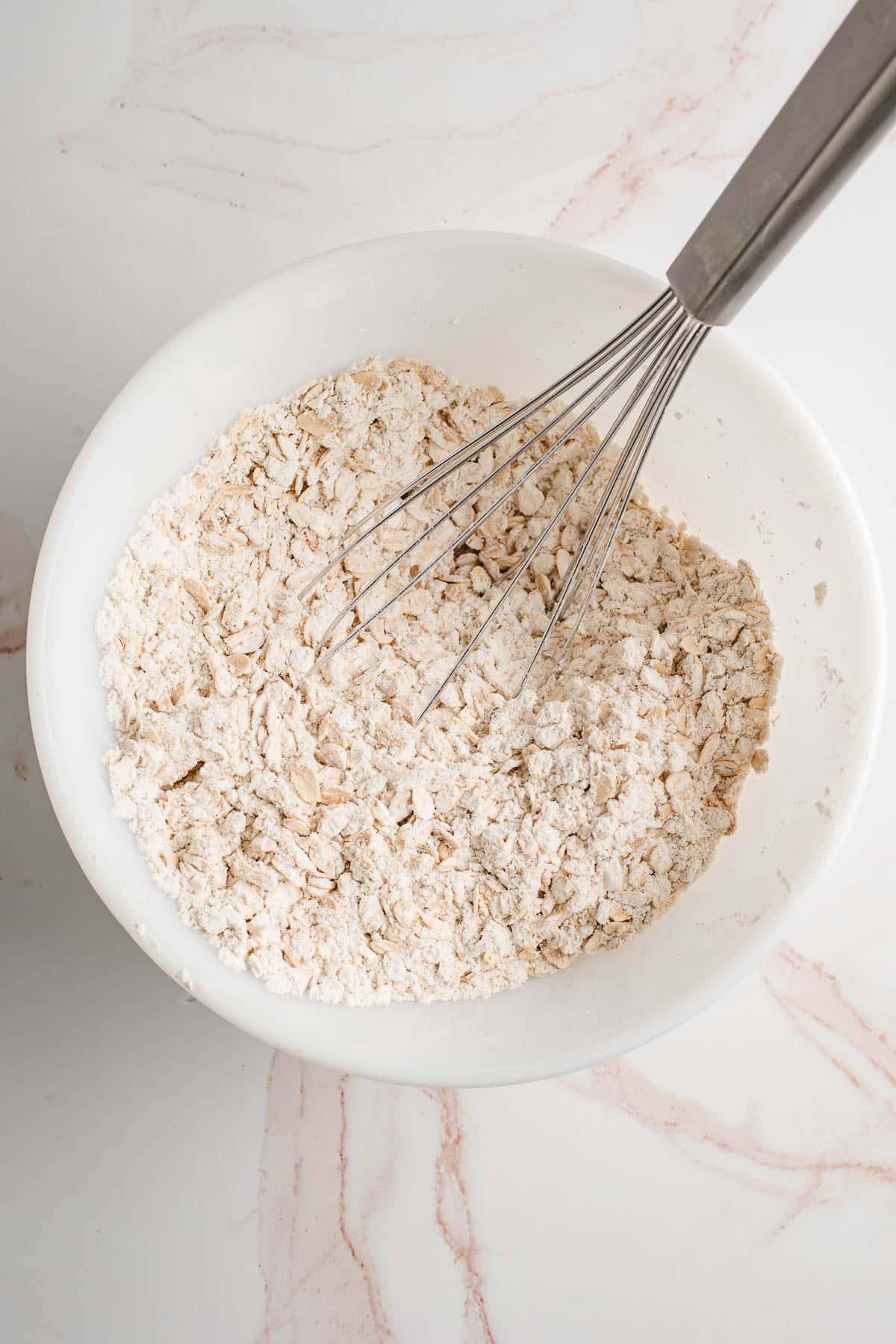 Rolled oats, flour, cinnamon, baking soda, and salt whisked in a white mixing bowl.