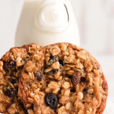 Two chewy oatmeal raisin cookies propped beside a small pitcher of milk.