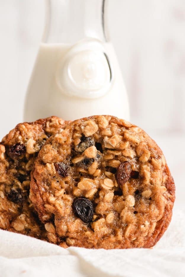 Two chewy oatmeal raisin cookies propped beside a small pitcher of milk.