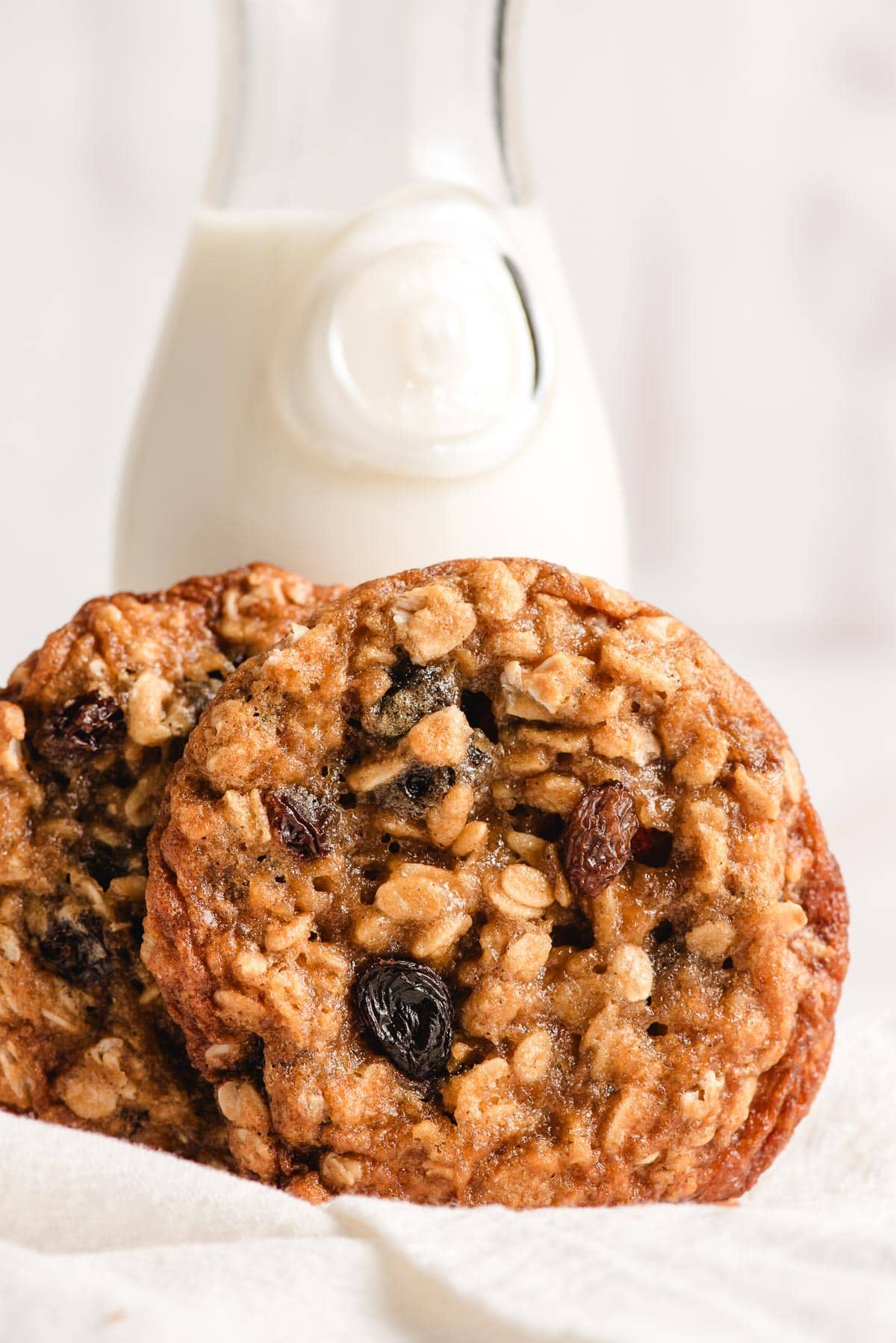 Two chewy oatmeal raisin cookies propped beside a small pitcher of milk.