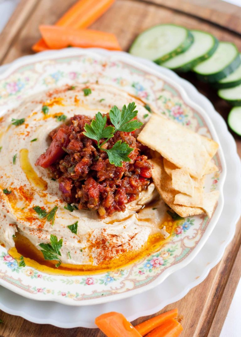 red pepper dip on top of hummus in a flowered bowl with pita chips resting on the side