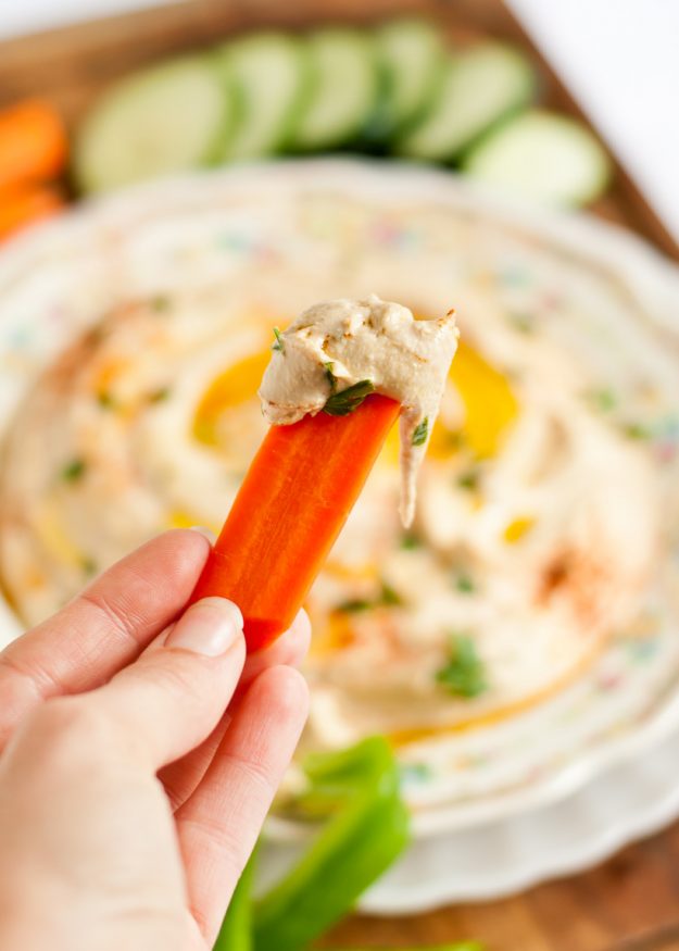 Image of a hand holding a carrot stick dipped in Homemade Hummus