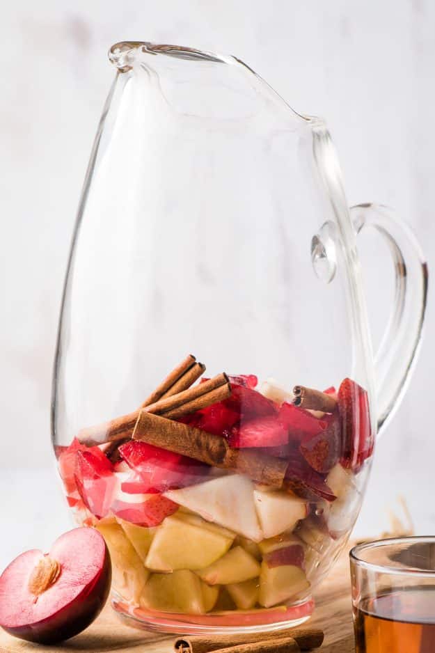 Large glass pitcher filled with apples, plums, pears, and cinnamon sticks.