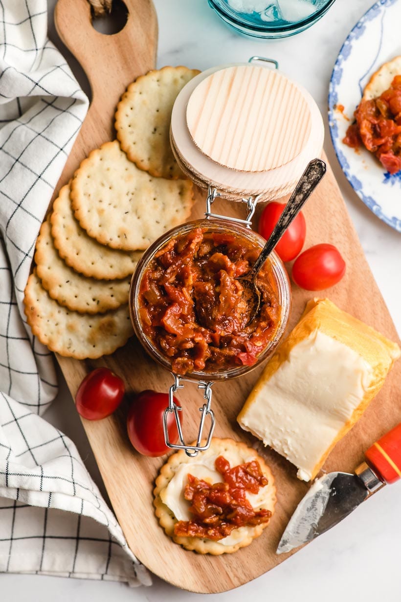 Wooden cutting board filled with crackers, cheese, and a jar of tomato bacon jam.