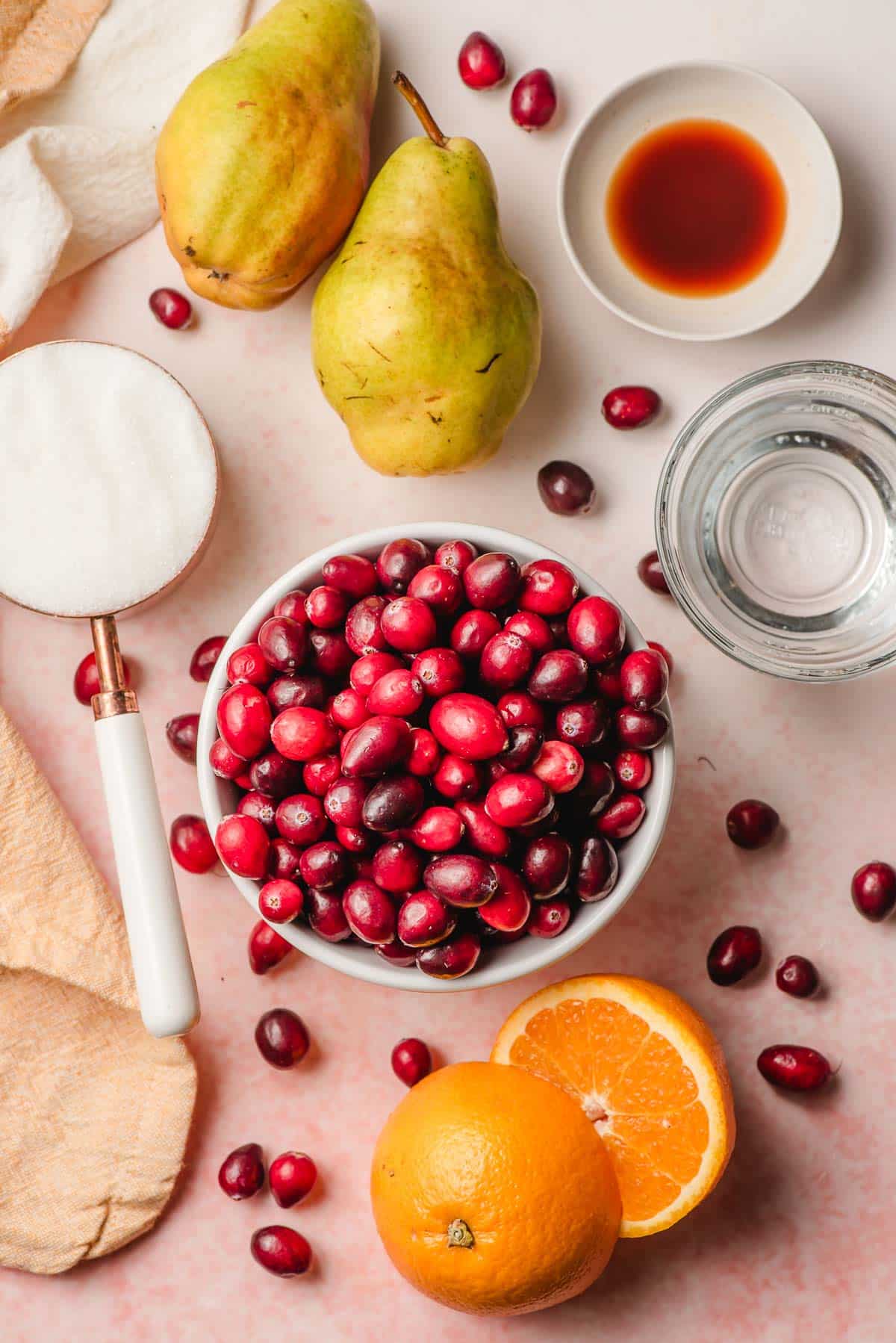 Bowl of cranberries, sliced orange, cup of sugar, water, vanilla, and pears on a blush pink background.