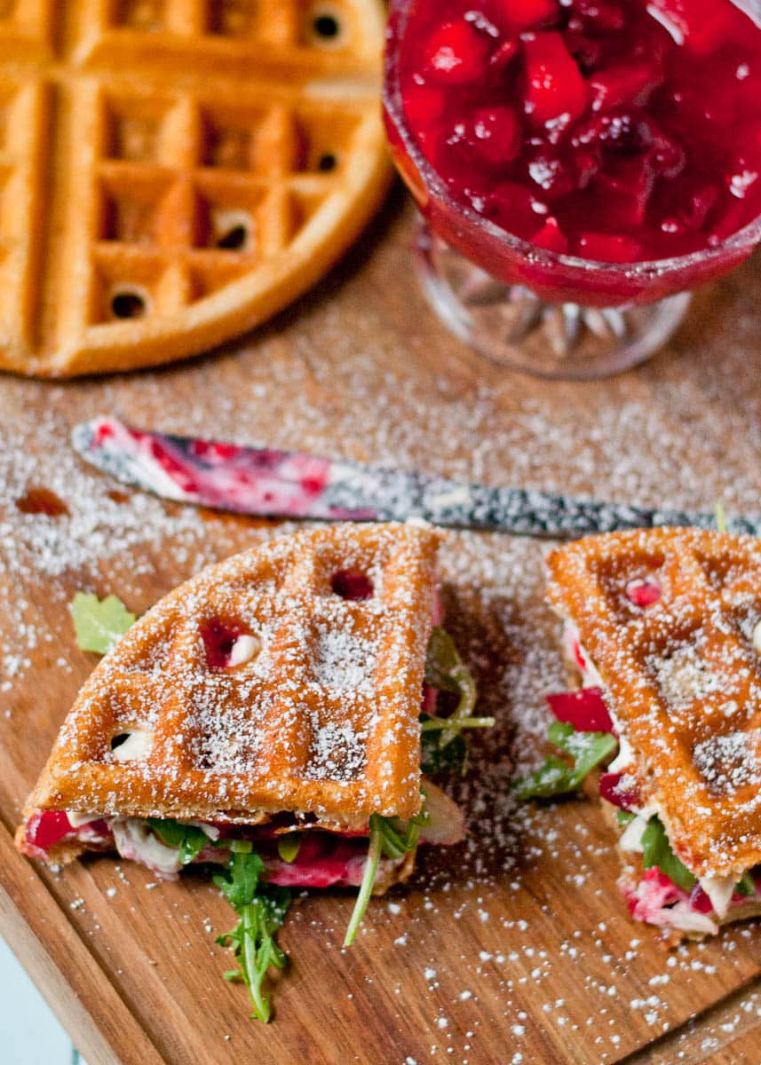 Two turkey and cranberry waffle sandwiches on a wooden cutting board with cranberry sauce in the back.