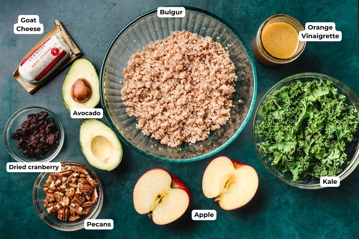 Bulgur wheat, a halved avocao, a halved apple, bowls of pecans and cranberries and kale, goat cheese, and orange vinaigrette on a dark background.
