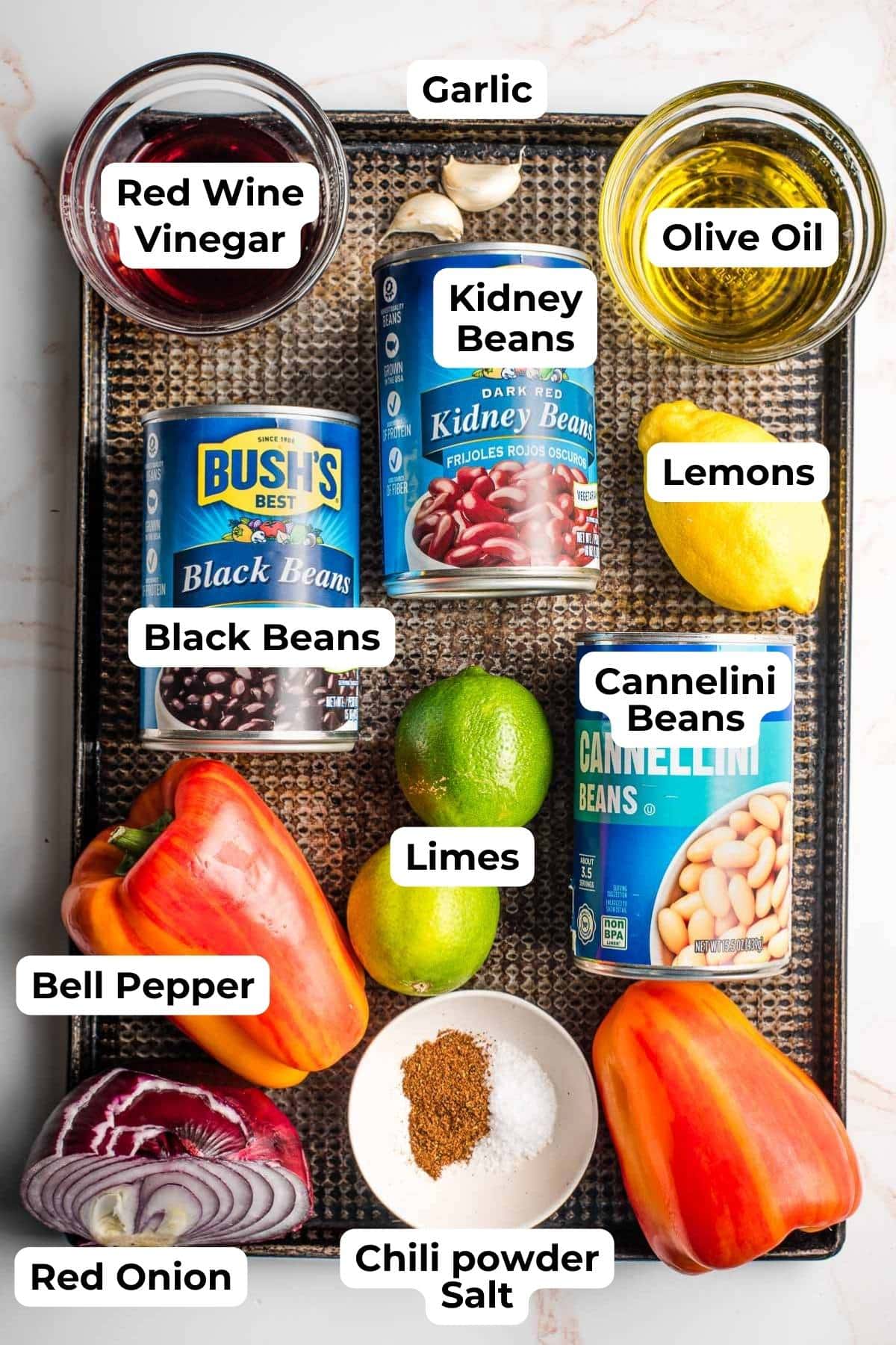 Red wine viengar, olive oil, three cans of beans, bell peppers, lemon, lime, red onion, and seasonings on a sheet pan.
