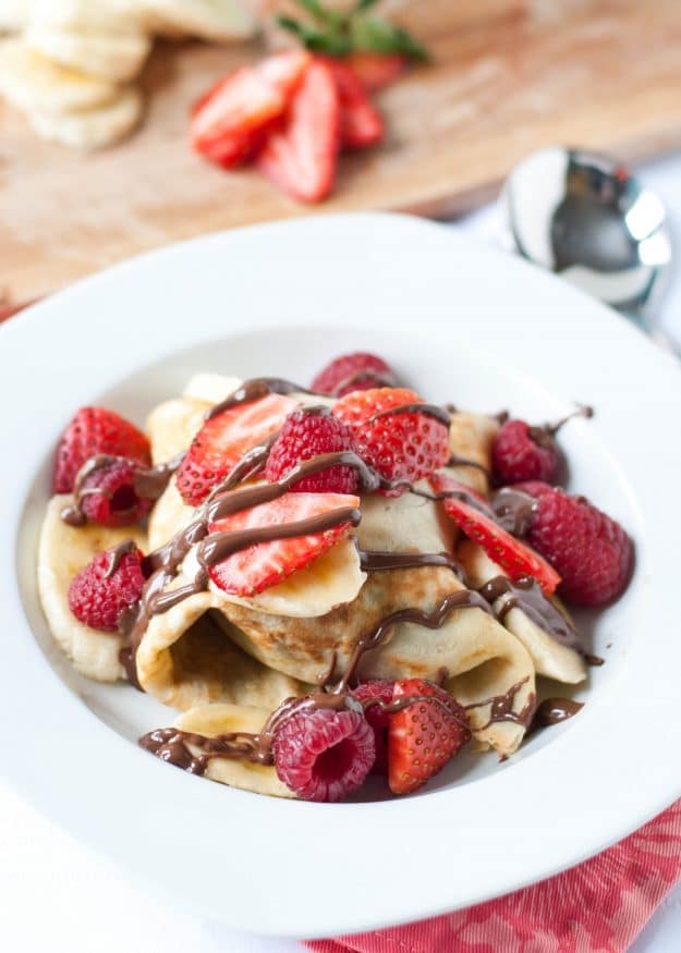 Crepes on a white plate topped with strawberries, bananas, and nutella