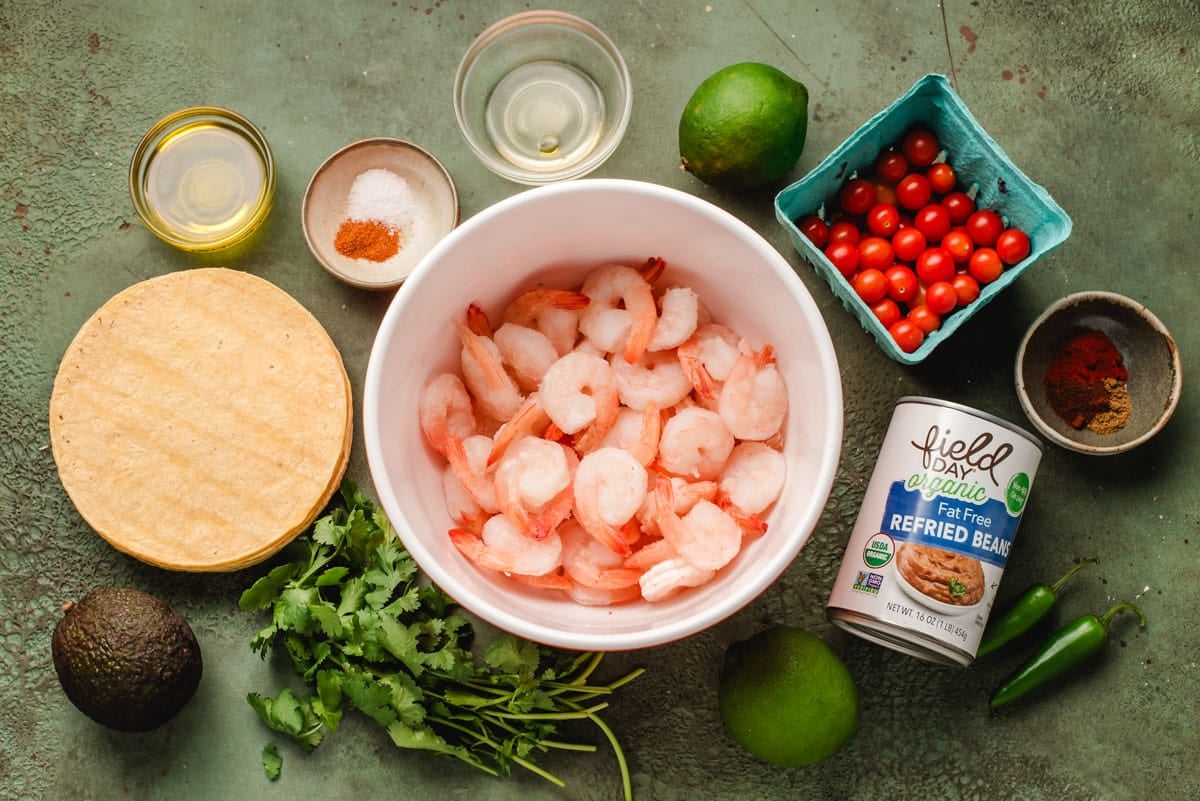 Corn tortillas, shrimp, avocado, cilantro, refried beans, jalapeno, tomatoes, tequila, seasonings, and limes on a green background.