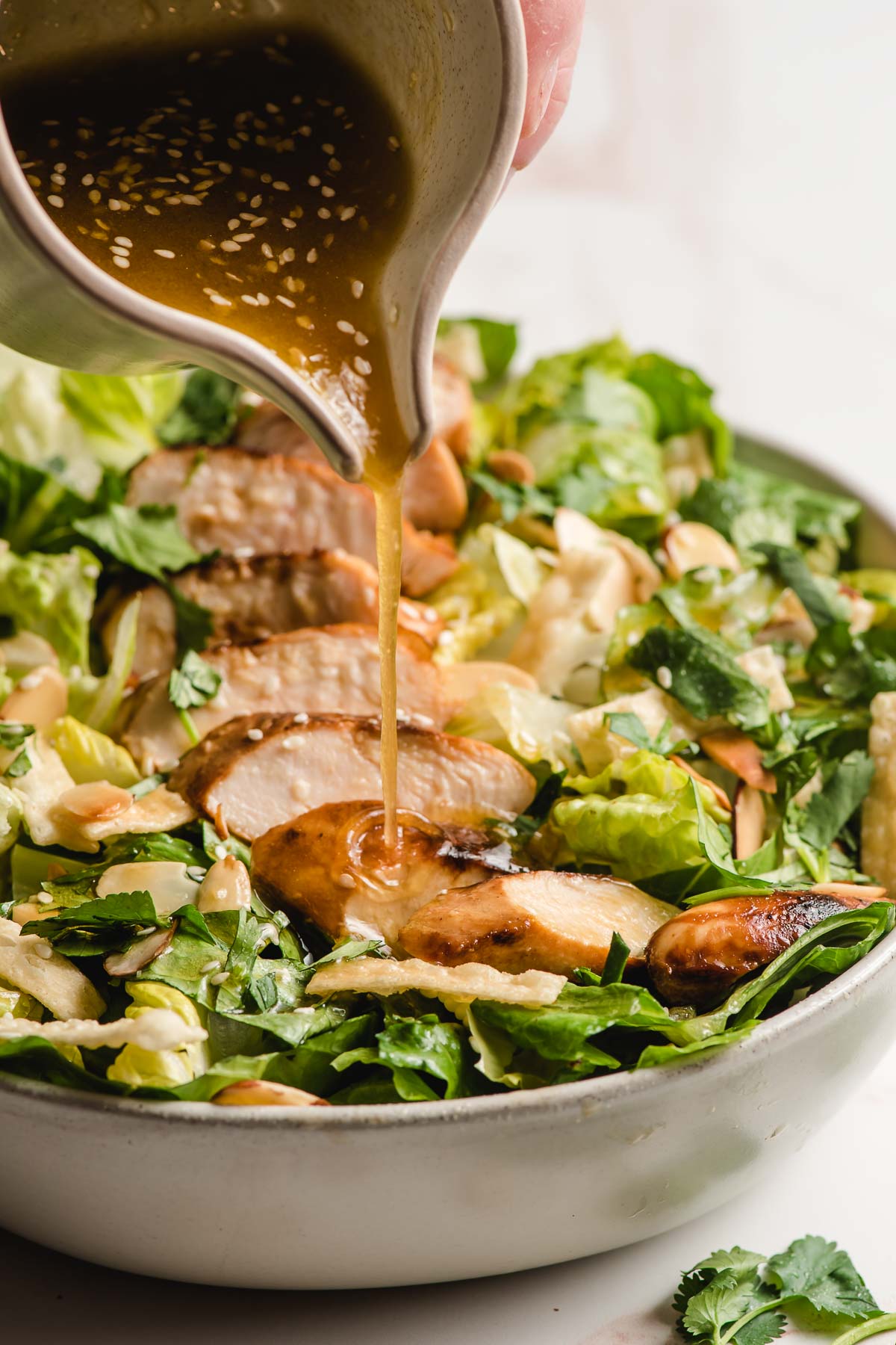 Asian sesame dressing being drizzled from a small pitcher onto a bowl of Asian chicken salad.