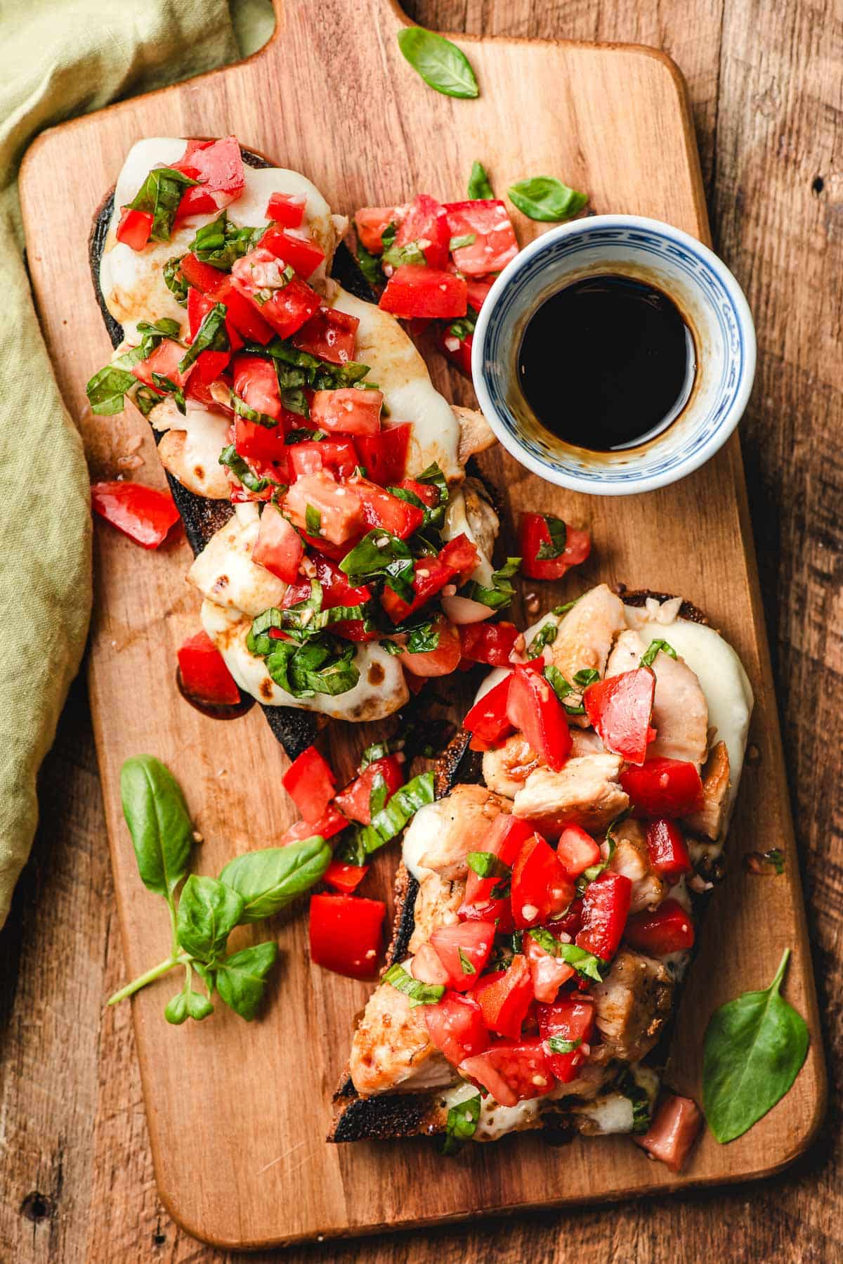 Two open face bruschetta chicken sandwiches on a wooden cutting board with a bowl of balsamic.