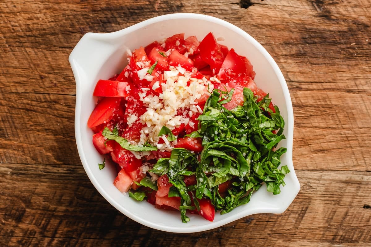 Chopped roma tomatoes, garlic, basil, and balsamic in a white bowl.