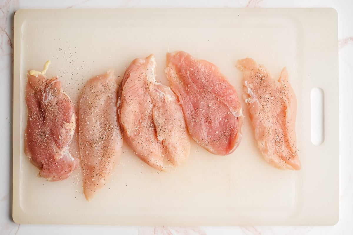 Butterflied chicken breasts seasoned with salt and pepper on a cutting board.
