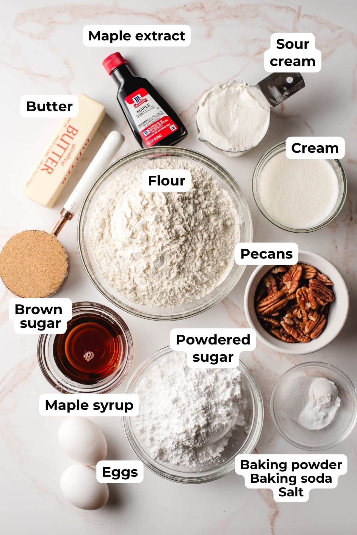 Baking staples, maple syrup, butter, brown sugar, powdered sugar, sour cream, cream, and eggs on a marble background.