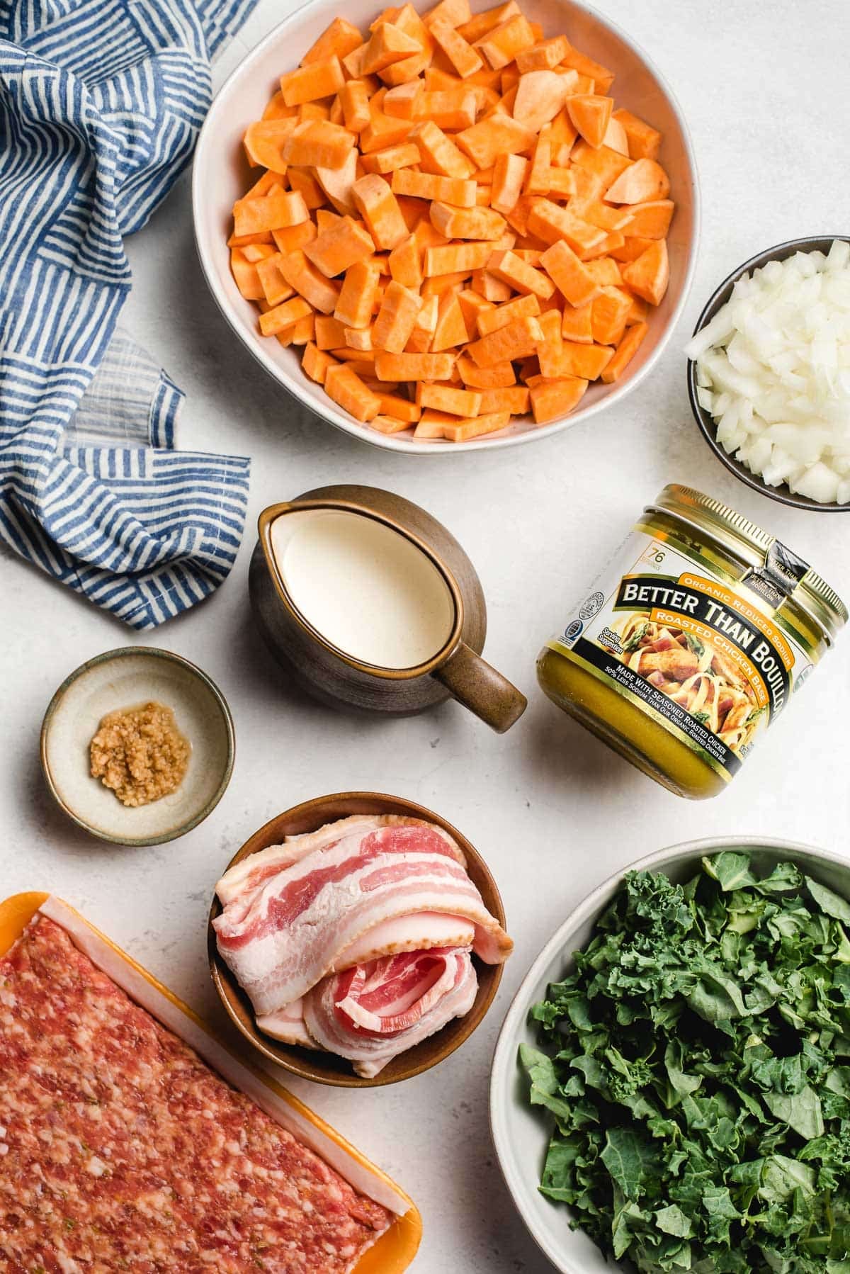Chopped sweet potatoes, cream, garlic, onion, bacon, sausage, chicken bouillon, and kale in bowls.
