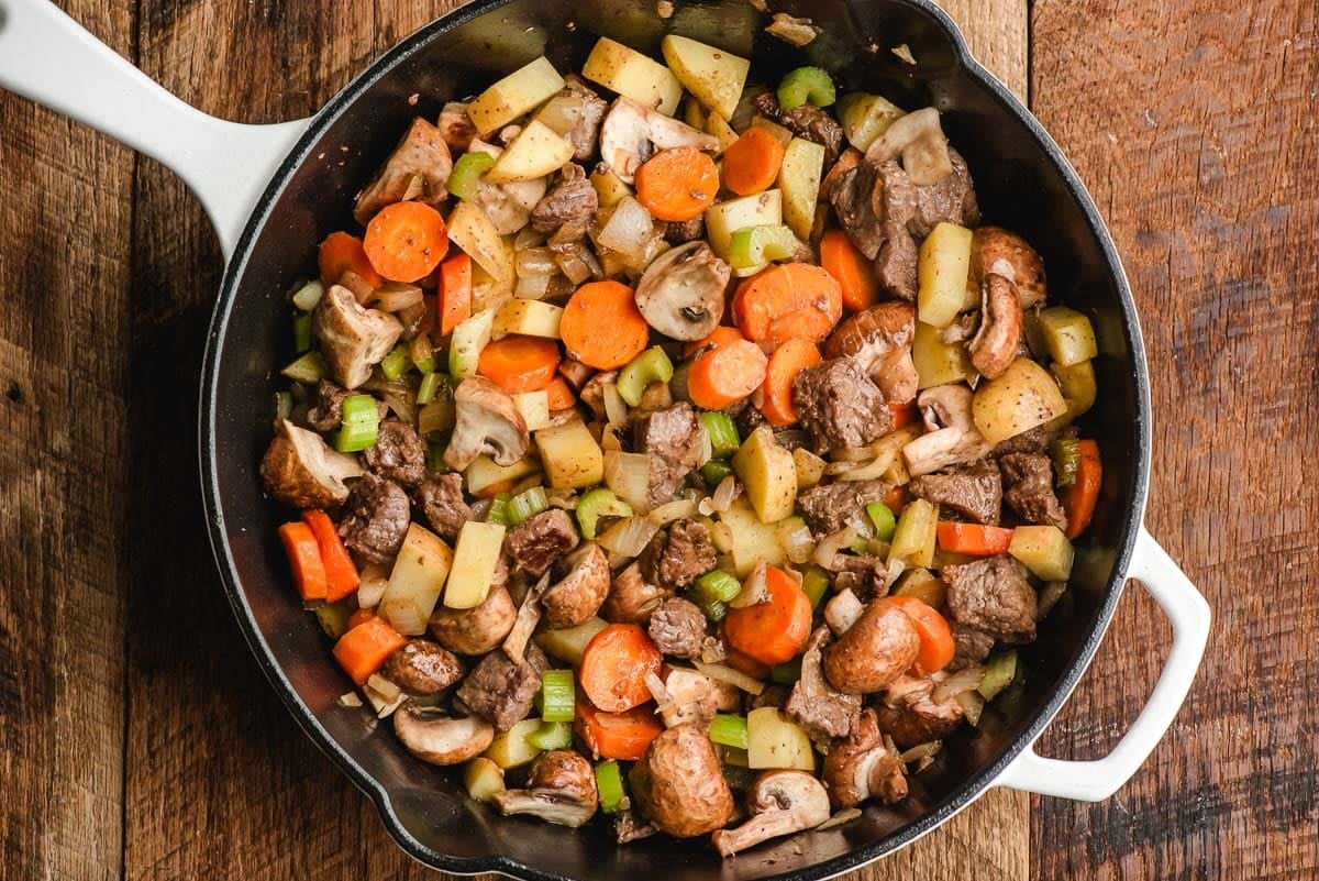 Stew beef, carrots, onions, celery, mushrooms, and potatoes in a cast iron skillet.
