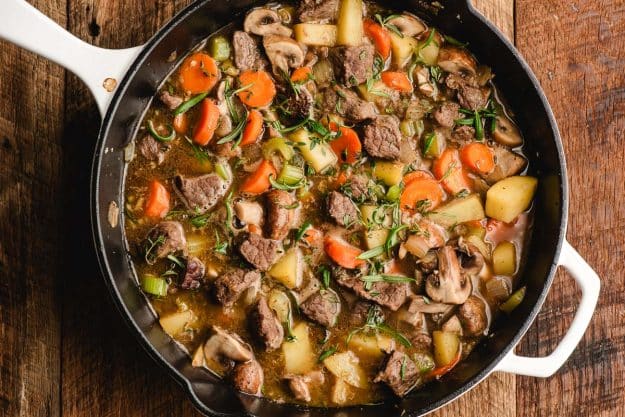 Guinness, beef broth, and fresh herbs with veggies and beef in a cast iron skillet.