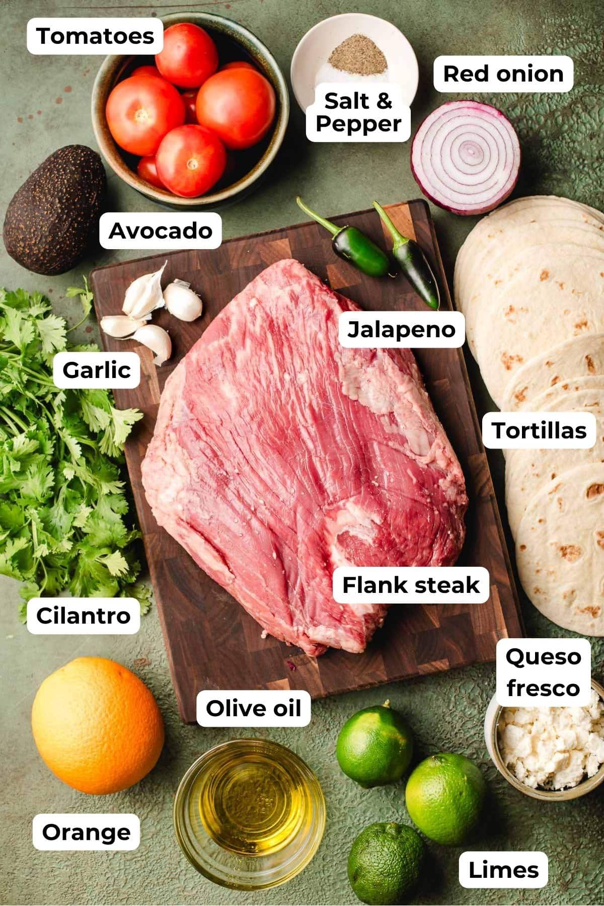 Flank steak on a cutting board, surrounded by cilantro, orange, limes, tomatoes, avocado, onion, queso fresco, and tortillas.